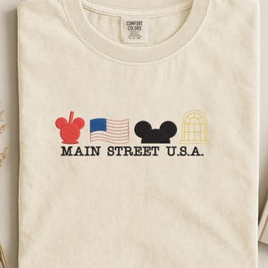 K&ouml;nnte beinhalten: Cremefarbenes T-Shirt mit gestickten Designs. Es zeigt einen roten Mickey Mouse-Kopf, eine blau-wei&szlig; amerikanische Flagge, einen schwarzen Mickey Mouse-Kopf und ein gelbes Bogenfenster. Der Text "MAIN STREET U.S.A." ist darunter gestickt.