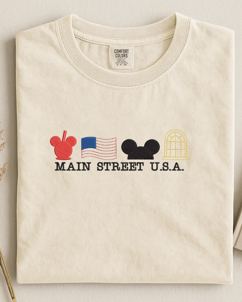 K&ouml;nnte beinhalten: Cremefarbenes T-Shirt mit gestickten Designs. Es zeigt einen roten Mickey Mouse-Kopf, eine blau-wei&szlig; amerikanische Flagge, einen schwarzen Mickey Mouse-Kopf und ein gelbes Bogenfenster. Der Text "MAIN STREET U.S.A." ist darunter gestickt.