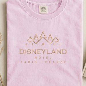 May include: A light pink t-shirt with a gold embroidered design featuring a crown and the words "DISNEYLAND HOTEL PARIS, FRANCE". The shirt is made of a soft material and has a crew neck.