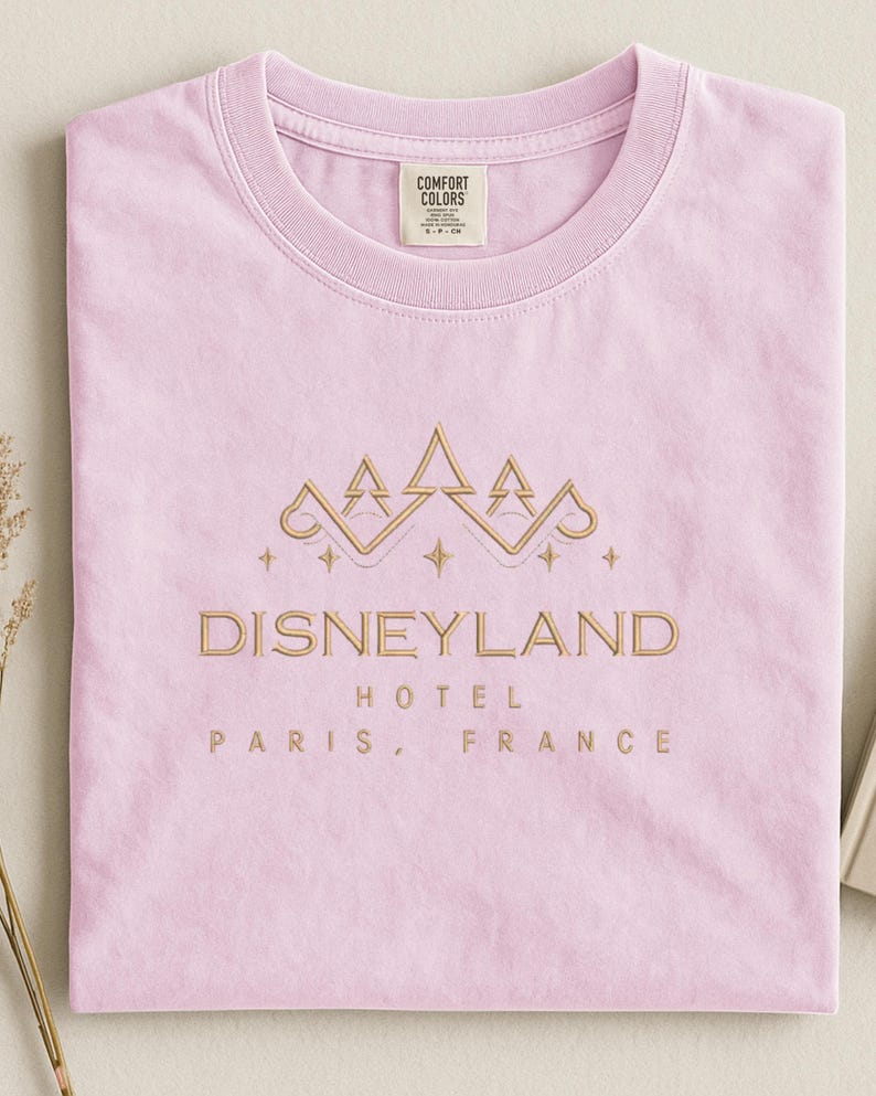 May include: A light pink t-shirt with a gold embroidered design featuring a crown and the words "DISNEYLAND HOTEL PARIS, FRANCE". The shirt is made of a soft material and has a crew neck.