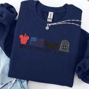 K&ouml;nnte beinhalten: Marineblaues Sweatshirt mit gesticktem Main Street U.S.A.-Design. Es zeigt rote, schwarze und goldfarbene Stickelemente, darunter ein Mickey Mouse-Kopf, eine amerikanische Flagge und ein Fenster. Eine silberne Halskette mit Sonnenanh&auml;nger liegt darauf.