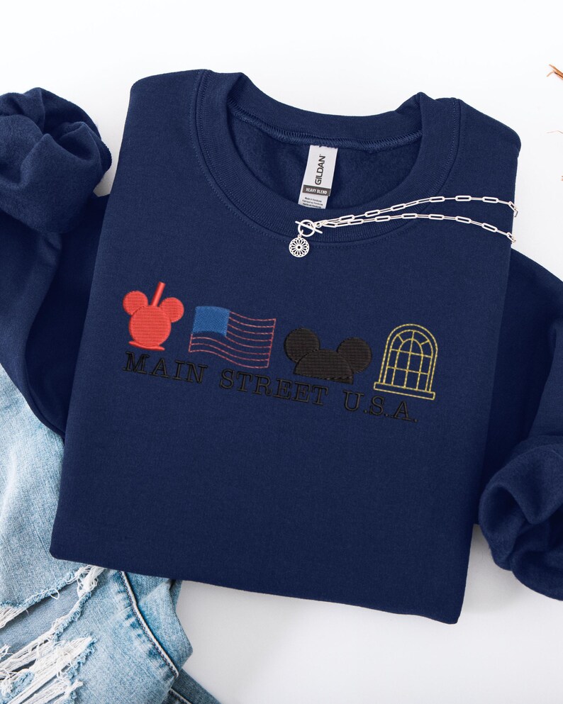 K&ouml;nnte beinhalten: Marineblaues Sweatshirt mit gesticktem Main Street U.S.A.-Design. Es zeigt rote, schwarze und goldfarbene Stickelemente, darunter ein Mickey Mouse-Kopf, eine amerikanische Flagge und ein Fenster. Eine silberne Halskette mit Sonnenanh&auml;nger liegt darauf.