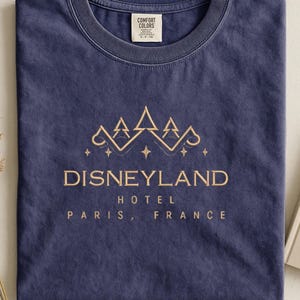 May include: A navy blue t-shirt with the words "Disneyland Hotel Paris, France" in gold lettering. The shirt features a gold graphic of stylized trees and stars above the text. The shirt is made of a soft, comfortable material.
