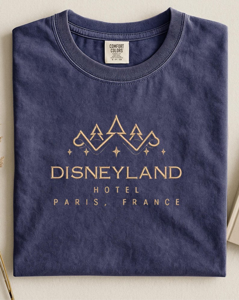 May include: A navy blue t-shirt with the words "Disneyland Hotel Paris, France" in gold lettering. The shirt features a gold graphic of stylized trees and stars above the text. The shirt is made of a soft, comfortable material.