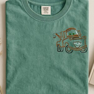 Puede incluir: Camiseta verde salvia con un dise&ntilde;o de dibujos animados de una gr&uacute;a en naranja y negro. La gr&uacute;a tiene ojos verdes y una boca sonriente. La camiseta tiene una etiqueta Comfort Colors.