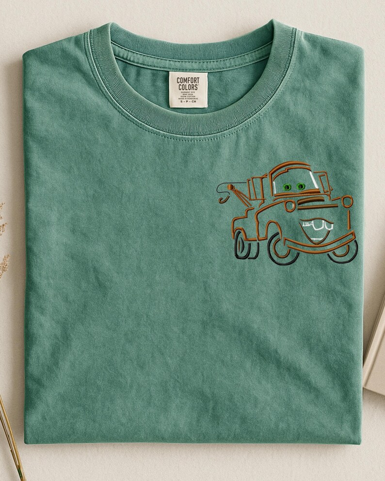 Puede incluir: Camiseta verde salvia con un dise&ntilde;o de dibujos animados de una gr&uacute;a en naranja y negro. La gr&uacute;a tiene ojos verdes y una boca sonriente. La camiseta tiene una etiqueta Comfort Colors.