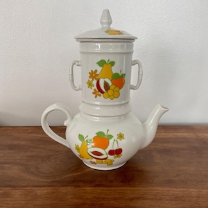 May include: White ceramic teapot with a fruit design. The teapot has a removable lid and a strainer. The fruit design features pears, apples, cherries, and peaches in shades of yellow, orange, red, and green.