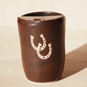 Puede incluir: Una taza de cer&aacute;mica marr&oacute;n con un dise&ntilde;o de herradura blanca. La taza tiene una forma curva y un peque&ntilde;o agujero en la parte superior. El dise&ntilde;o de la herradura est&aacute; centrado en la parte delantera de la taza.
