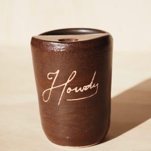May include: A handcrafted, brown ceramic cup with the word "Howdy" in a cursive font. The cup has a rounded base and a slightly flared top. The background is a light wood surface.