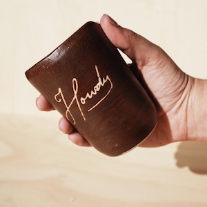 May include: A brown ceramic mug with the word "Howdy" in a cursive font. The mug has a slightly tapered shape and is held by a person's hand. The background is a light wood surface.