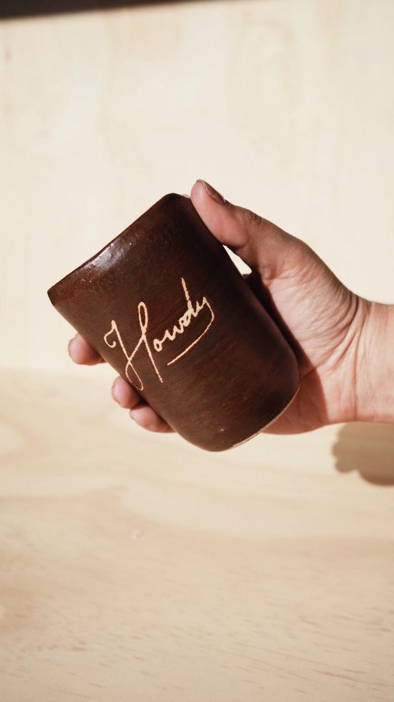 May include: A brown ceramic mug with the word "Howdy" in a cursive font. The mug has a slightly tapered shape and is held by a person's hand. The background is a light wood surface.