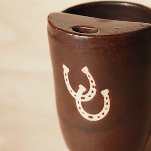 Puede incluir: Una taza de cer&aacute;mica marr&oacute;n con un dise&ntilde;o de herradura blanca. La taza tiene forma redondeada con la parte superior plana y un peque&ntilde;o agujero. El dise&ntilde;o de la herradura est&aacute; en la parte delantera.