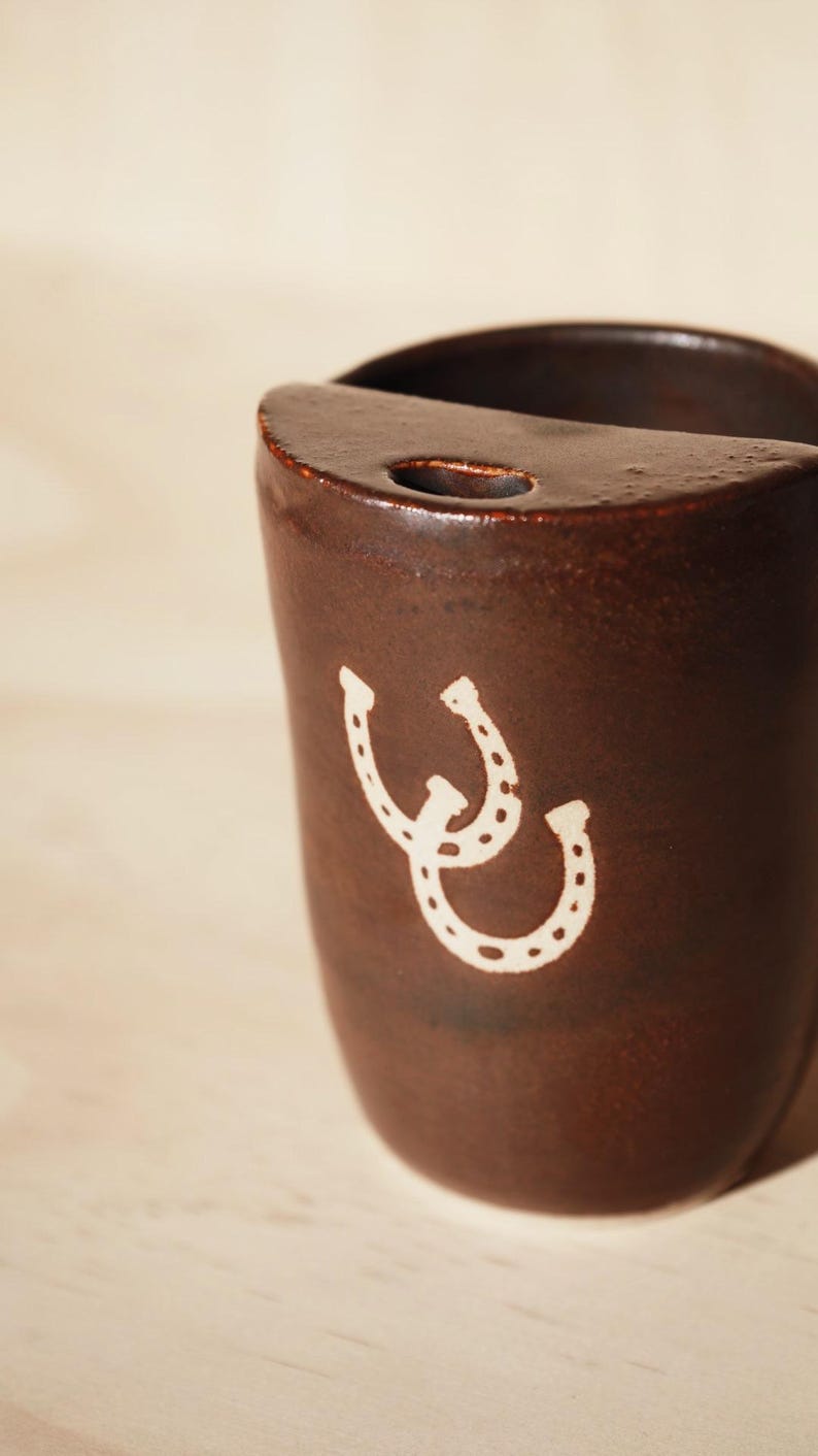 Puede incluir: Una taza de cer&aacute;mica marr&oacute;n con un dise&ntilde;o de herradura blanca. La taza tiene forma redondeada con la parte superior plana y un peque&ntilde;o agujero. El dise&ntilde;o de la herradura est&aacute; en la parte delantera.