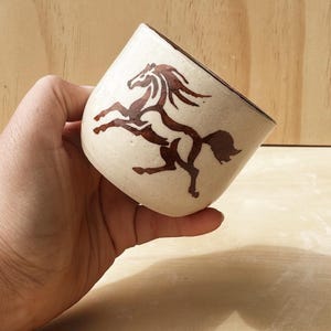 May include: Cream-colored ceramic cup with a brown horse illustration. The horse is depicted mid-gallop. The rim of the cup is brown. The cup is held in a hand against a light wood background.