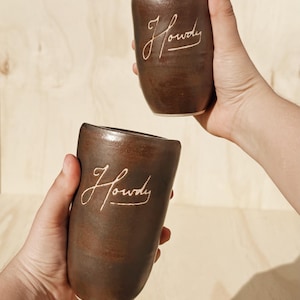 May include: Two brown ceramic mugs with the word "Howdy" in a cursive font. The mugs are held up against a light brown wooden background. The mugs have a smooth, rounded shape.