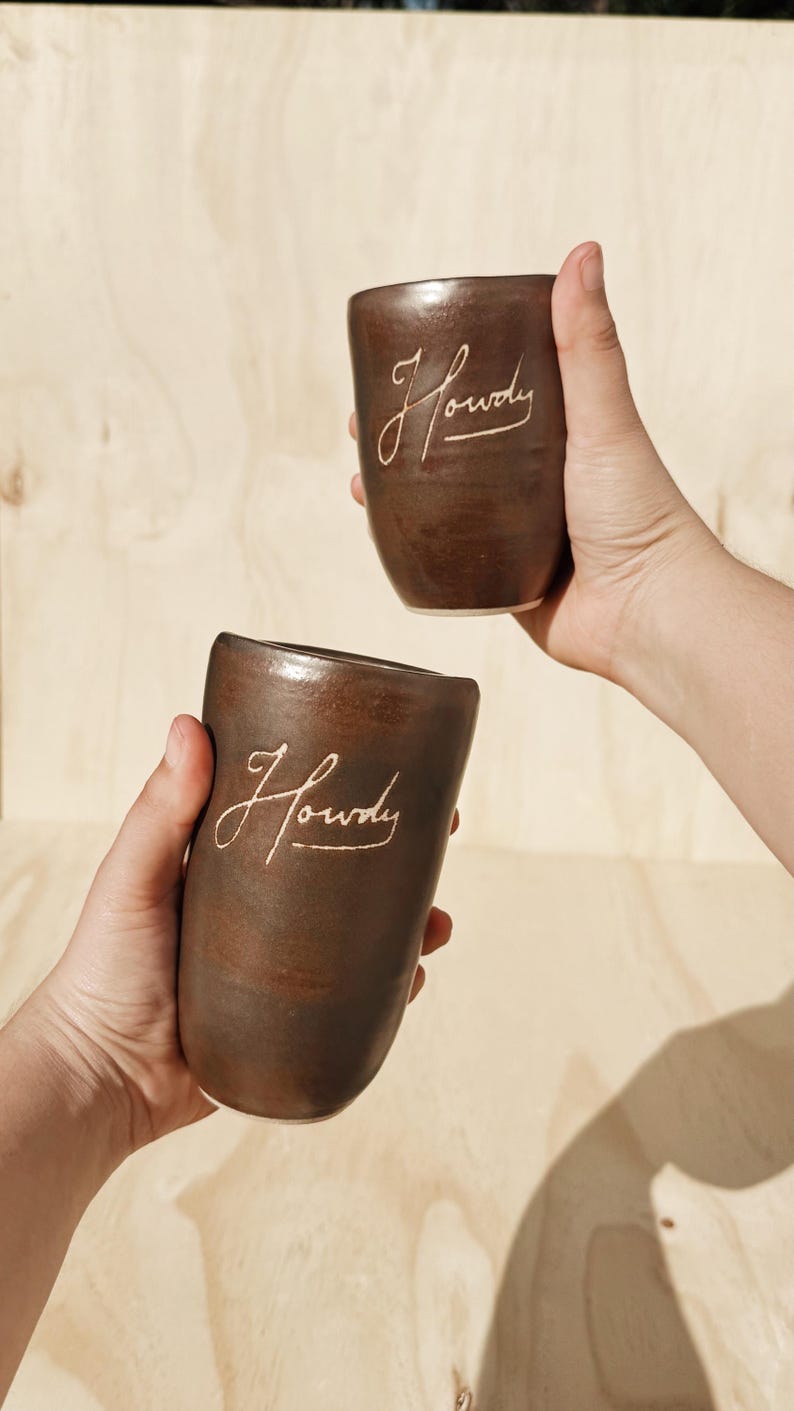 May include: Two brown ceramic mugs with the word "Howdy" in a cursive font. The mugs are held up against a light brown wooden background. The mugs have a smooth, rounded shape.