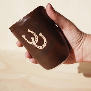 Puede incluir: Una taza de cer&aacute;mica marr&oacute;n con un dise&ntilde;o de herradura blanca. La taza se sostiene en una mano y tiene una forma ligeramente c&oacute;nica. El dise&ntilde;o de la herradura est&aacute; centrado en el lateral de la taza.