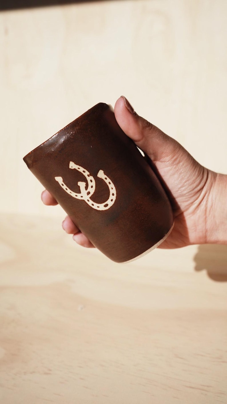 Puede incluir: Una taza de cer&aacute;mica marr&oacute;n con un dise&ntilde;o de herradura blanca. La taza se sostiene en una mano y tiene una forma ligeramente c&oacute;nica. El dise&ntilde;o de la herradura est&aacute; centrado en el lateral de la taza.