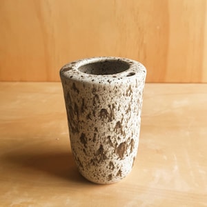 May include: A cylindrical ceramic vessel featuring a speckled off-white base with dark brown, irregular paint splatters across the exterior surface. The rim is rounded and smooth, and the interior is hollow with a small circular opening near the top edge.