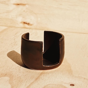 May include: A dark brown ceramic napkin ring with a smooth, glossy finish and a U-shaped cutout design. The object features a rounded, cylindrical form with a wide vertical opening on one side, showcasing a deep, uniform dark brown glaze.