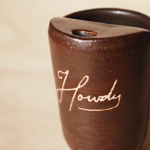 May include: A brown ceramic mug with the word "Howdy" in a cursive font. The mug has a rounded top with a small hole and a smooth, glossy finish. The background is a light wood surface.