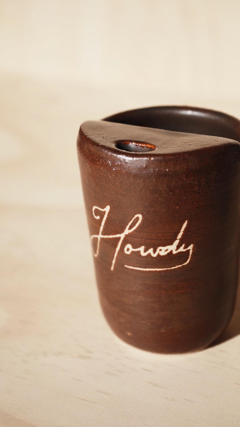 May include: A brown ceramic mug with the word "Howdy" in a cursive font. The mug has a rounded top with a small hole and a smooth, glossy finish. The background is a light wood surface.