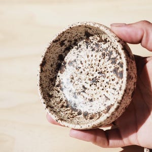 May include: A small, speckled ceramic bowl with a cream-colored base and dark brown spots. The bowl is held in a hand, showcasing its rounded shape and textured surface. The interior has a speckled pattern.