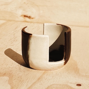 May include: A small, handcrafted ceramic bowl with a unique design. The bowl features a two-tone color scheme, with a dark brown exterior and a cream-colored interior. The bowl is approximately 2 inches tall and 3 inches wide.