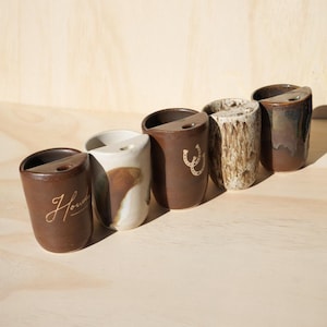 Puede incluir: Una colecci&oacute;n de seis tazas de cer&aacute;mica hechas a mano en varios estilos y colores. Las tazas son marrones, blancas y texturizadas, algunas con un dise&ntilde;o de herradura o la palabra "Howdy". Cada taza tiene una forma y un acabado &uacute;nicos.