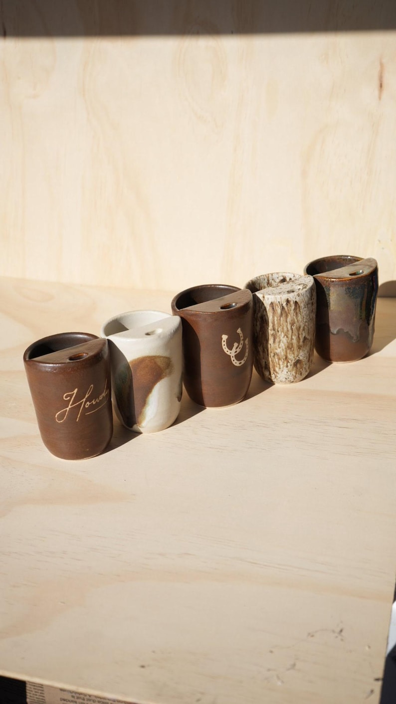 Puede incluir: Una colecci&oacute;n de seis tazas de cer&aacute;mica hechas a mano en varios estilos y colores. Las tazas son marrones, blancas y texturizadas, algunas con un dise&ntilde;o de herradura o la palabra "Howdy". Cada taza tiene una forma y un acabado &uacute;nicos.