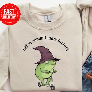 May include: Beige sweatshirt with the phrase "Off to commit mom foolery" in black. A green frog wearing a purple witch hat is riding a scooter. A red "Fast Delivery" sticker is in the upper left corner.