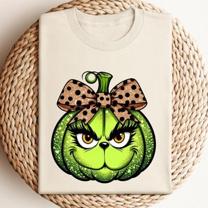 May include: A white t-shirt with a green pumpkin with a brown and white polka dot bow. The pumpkin has a green face with black eyelashes, orange eyes, and a black nose.