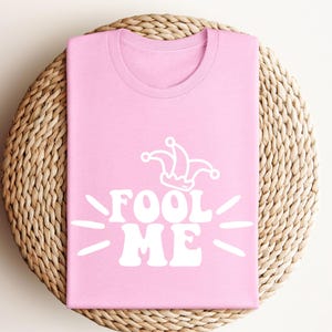 Puede incluir: Camiseta rosa claro con la frase blanca "FOOL ME" y un gorro de buf&oacute;n. La camiseta est&aacute; doblada sobre una superficie redonda de mimbre. El dise&ntilde;o es sencillo y llamativo.