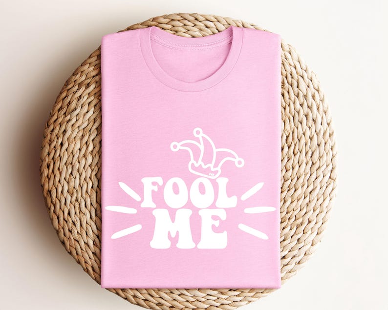 Puede incluir: Camiseta rosa claro con la frase blanca "FOOL ME" y un gorro de buf&oacute;n. La camiseta est&aacute; doblada sobre una superficie redonda de mimbre. El dise&ntilde;o es sencillo y llamativo.
