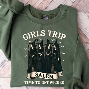 May include: Green sweatshirt with the text "GIRLS TRIP SALEM TIME TO GET WICKED." The graphic features four witches in black dresses and hats, with bats flying above them. The sweatshirt is a solid green color.