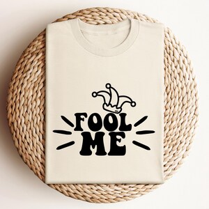 Puede incluir: Una camiseta color crema con las palabras "FOOL ME" en letras negras en negrita, acompa&ntilde;ada de un gr&aacute;fico de sombrero de buf&oacute;n. El dise&ntilde;o incluye l&iacute;neas radiantes, creando un visual l&uacute;dico. La camiseta se muestra sobre una superficie circular tejida.