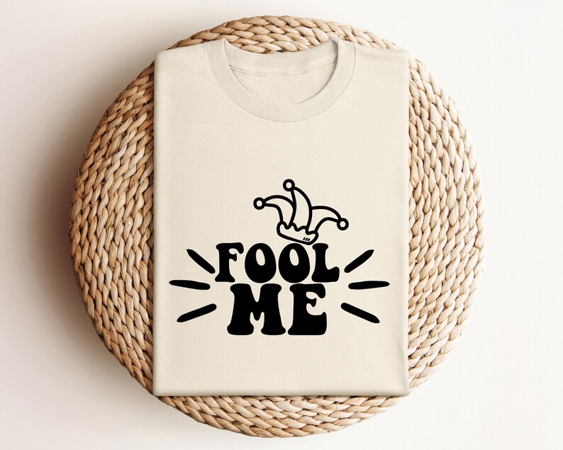 Puede incluir: Una camiseta color crema con las palabras "FOOL ME" en letras negras en negrita, acompa&ntilde;ada de un gr&aacute;fico de sombrero de buf&oacute;n. El dise&ntilde;o incluye l&iacute;neas radiantes, creando un visual l&uacute;dico. La camiseta se muestra sobre una superficie circular tejida.