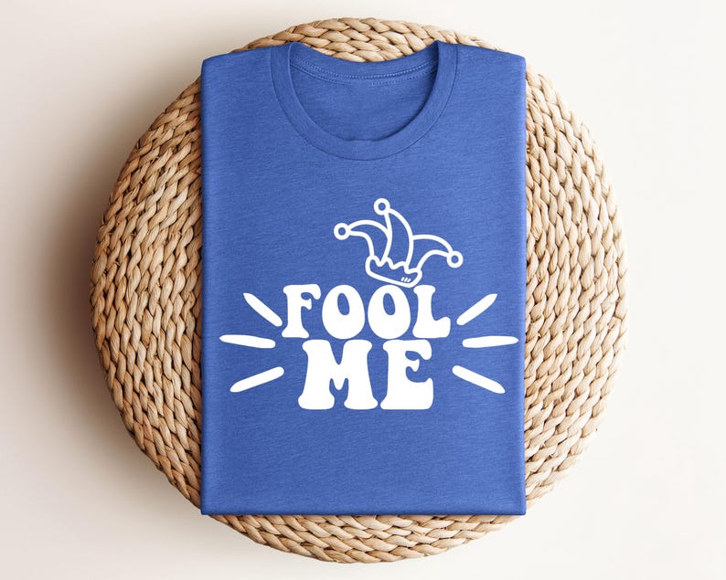Puede incluir: Una camiseta azul con la frase "FOOL ME" en blanco y un gorro de buf&oacute;n blanco. La camiseta est&aacute; doblada y colocada sobre un salvamanteles redondo y tejido. El dise&ntilde;o es sencillo y atrevido.