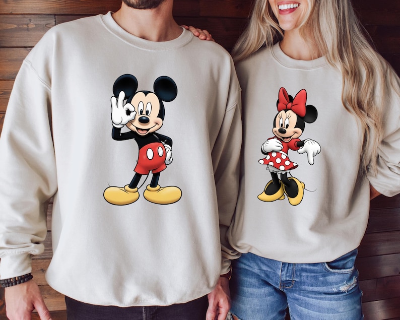 May include: Beige sweatshirts featuring Mickey Mouse and Minnie Mouse. Mickey is in red shorts and yellow shoes, giving a hand gesture. Minnie is in a red polka dot dress and bow. The sweatshirts are a neutral color.