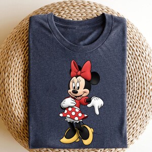 May include: A navy blue t-shirt featuring a colorful graphic of Minnie Mouse. Minnie is wearing a red dress with white polka dots, a red bow, white gloves, and yellow shoes. The shirt is folded on a woven surface.