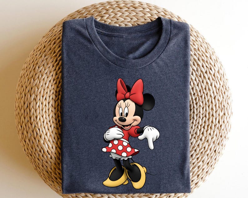 May include: A navy blue t-shirt featuring a colorful graphic of Minnie Mouse. Minnie is wearing a red dress with white polka dots, a red bow, white gloves, and yellow shoes. The shirt is folded on a woven surface.