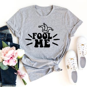 Puede incluir: Una camiseta gris con el texto negro "FOOL ME" y un gr&aacute;fico de sombrero de buf&oacute;n. La imagen incluye vaqueros azules, zapatillas blancas y un ramo de flores rosas y blancas.