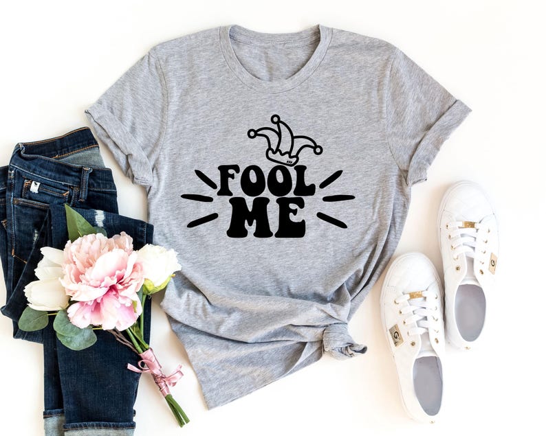 Puede incluir: Una camiseta gris con el texto negro "FOOL ME" y un gr&aacute;fico de sombrero de buf&oacute;n. La imagen incluye vaqueros azules, zapatillas blancas y un ramo de flores rosas y blancas.
