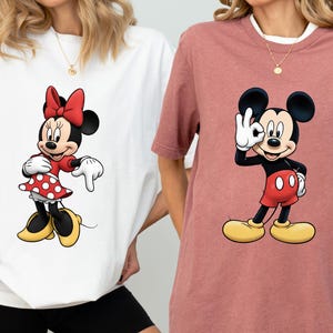 May include: Two t-shirts featuring cartoon characters. One is white with Minnie Mouse in a red dress with white polka dots and a red bow. The other is a mauve shirt with Mickey Mouse giving an "okay" hand gesture.