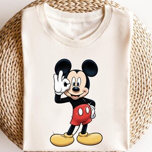 May include: Cream-colored t-shirt featuring a cartoon illustration of Mickey Mouse. Mickey is depicted in his classic attire: black body, red shorts, yellow shoes, and white gloves. He is making an "OK" hand gesture.
