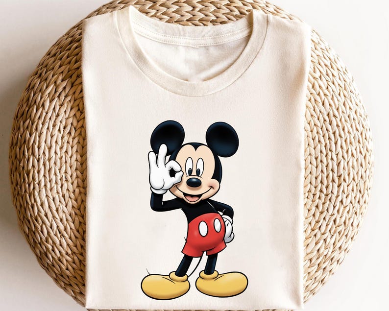 May include: Cream-colored t-shirt featuring a cartoon illustration of Mickey Mouse. Mickey is depicted in his classic attire: black body, red shorts, yellow shoes, and white gloves. He is making an "OK" hand gesture.