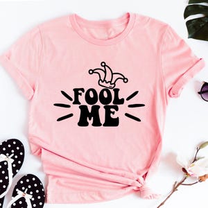 Puede incluir: Camiseta rosa claro con la frase "FOOL ME" en negro y un gorro de buf&oacute;n. La imagen incluye chanclas de lunares blancos y negros y gafas de sol.