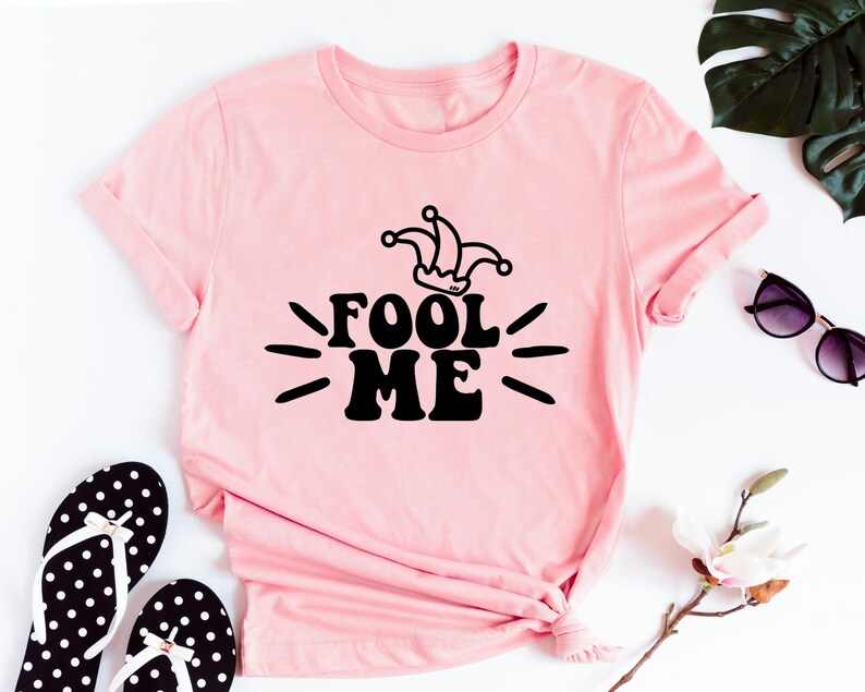 Puede incluir: Camiseta rosa claro con la frase "FOOL ME" en negro y un gorro de buf&oacute;n. La imagen incluye chanclas de lunares blancos y negros y gafas de sol.