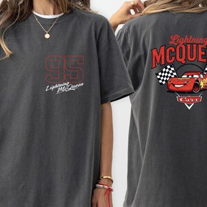 Puede incluir: Camiseta gris oscuro con el n&uacute;mero "95" en rojo y el texto "Lightning McQueen". La parte trasera de la camiseta muestra un gr&aacute;fico de Rayo McQueen con banderas a cuadros y el texto "Lightning McQueen Cars".