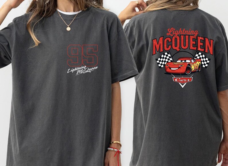 Puede incluir: Camiseta gris oscuro con el n&uacute;mero "95" en rojo y el texto "Lightning McQueen". La parte trasera de la camiseta muestra un gr&aacute;fico de Rayo McQueen con banderas a cuadros y el texto "Lightning McQueen Cars".